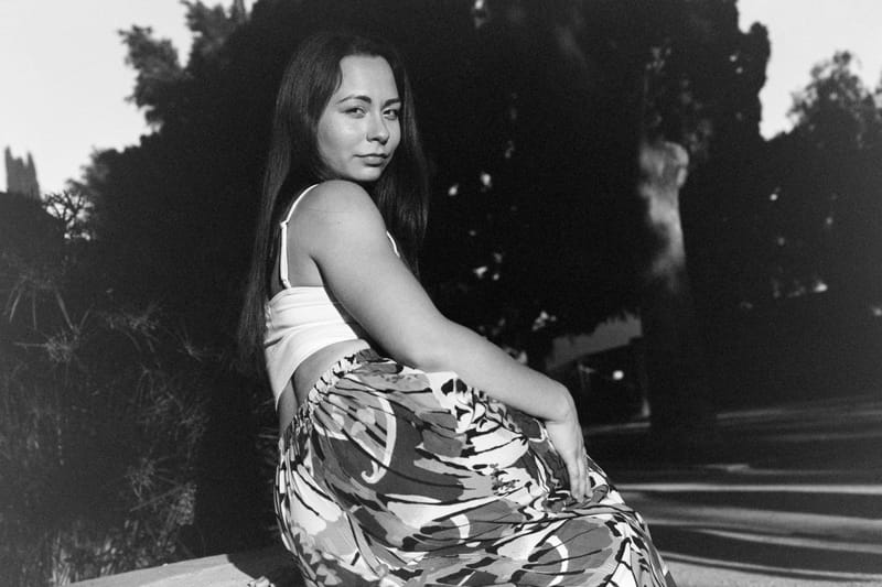 A young woman with long dark hair glances over her shoulder, seated outdoors in low golden light; black-and-white, dense foliage behind her in deep shadow.
