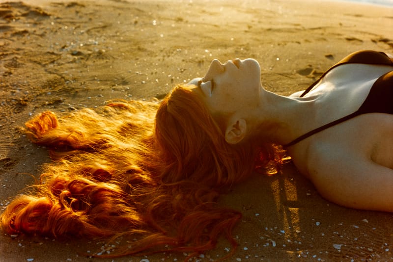 Woman lying on wet sand with head tilted back, her long copper hair fanned out and backlit by low golden sunset light.