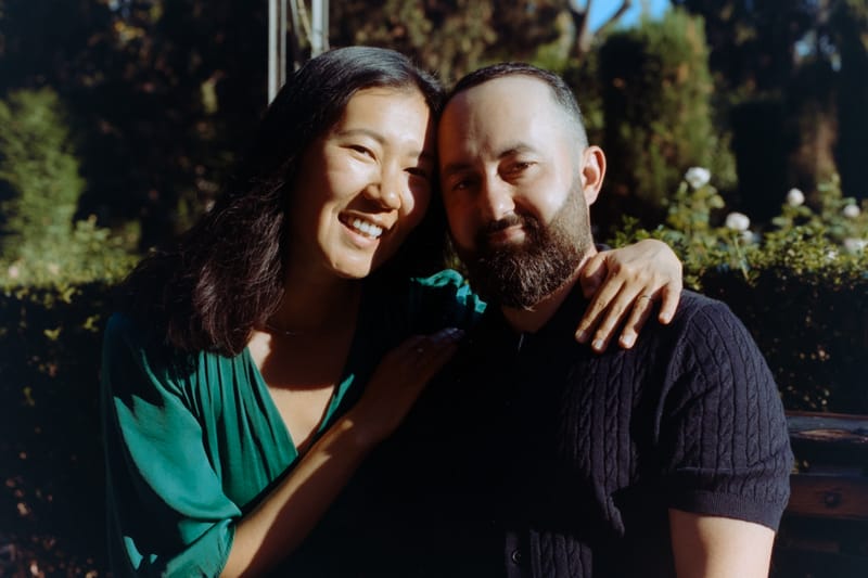 A couple leans head-to-head in dappled afternoon sun, her arm draped over his shoulder, flowering hedge behind them.