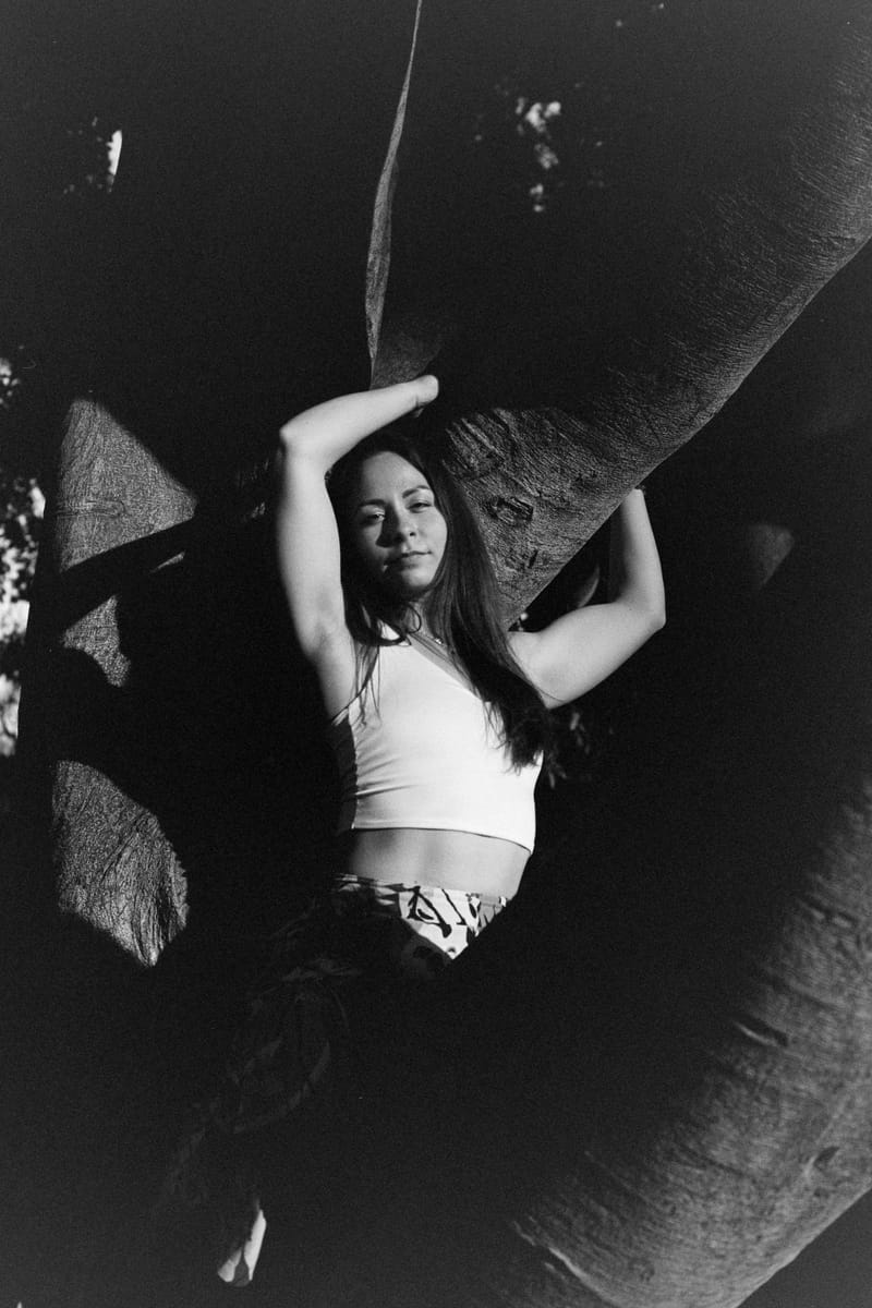 A young woman in a white crop top reclines against a tree trunk, arms raised overhead, her face and torso lit by a hard shaft of sunlight while deep black shadow swallows the surrounding bark.