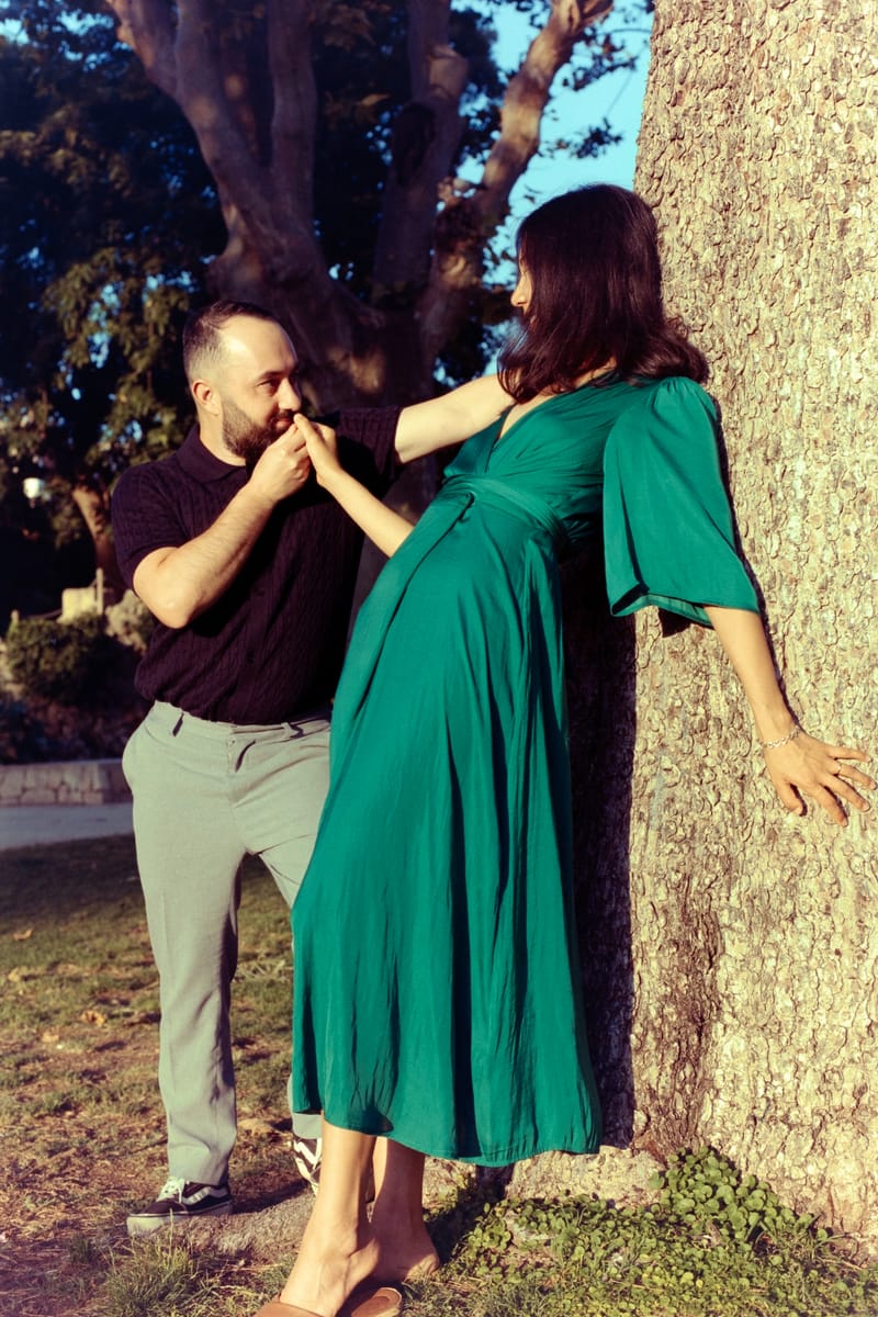 A bearded man in black kisses the hand of a woman in an emerald dress leaning against a sunlit tree trunk in a park.