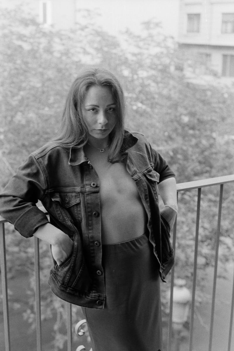 Woman in an open denim jacket leans on a balcony rail, gazing at the camera; blurred trees fill the soft gray light.