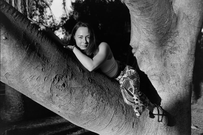 Black-and-white portrait of a young woman lying along a massive smooth tree limb, chin resting on her arms, patterned pants and strappy sandals trailing behind her as low sun rakes across the bark.