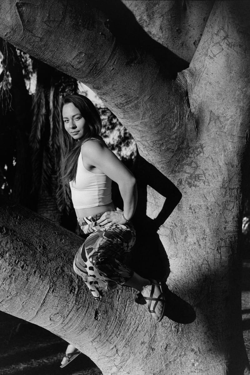A woman in a white crop top and camo pants reclines in the fork of a massive tree, framed by sunlit bark, in black and white.