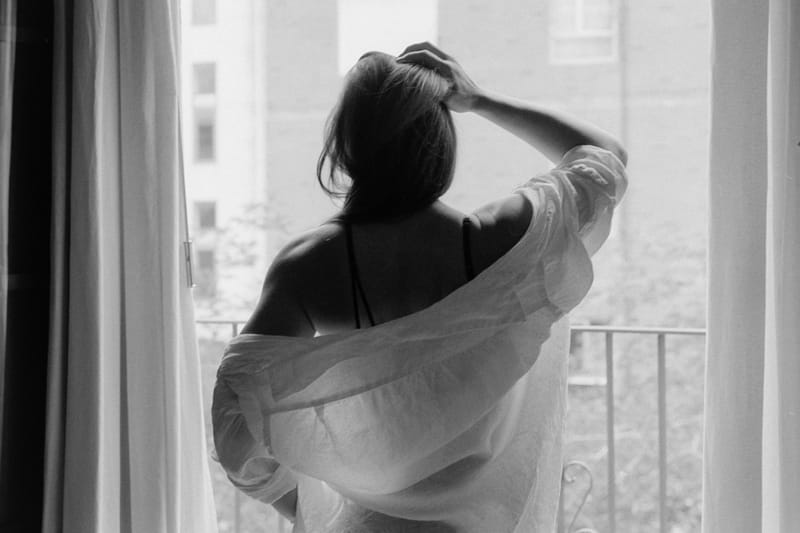 Backlit silhouette of a woman seen from behind, lifting her hair, an oversized linen shirt slipping off one shoulder as she stands at an open balcony window in soft, blown-out daylight.
