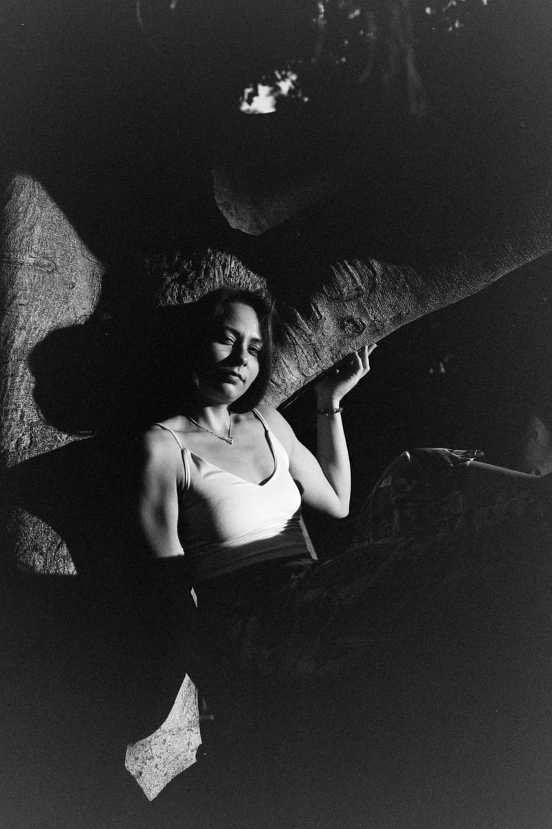 A woman in a white tank top reclines against a sunlit tree trunk, her face and shoulders caught in a narrow shaft of light while deep black shadow swallows the rest of the frame in this high-contrast monochrome portrait.