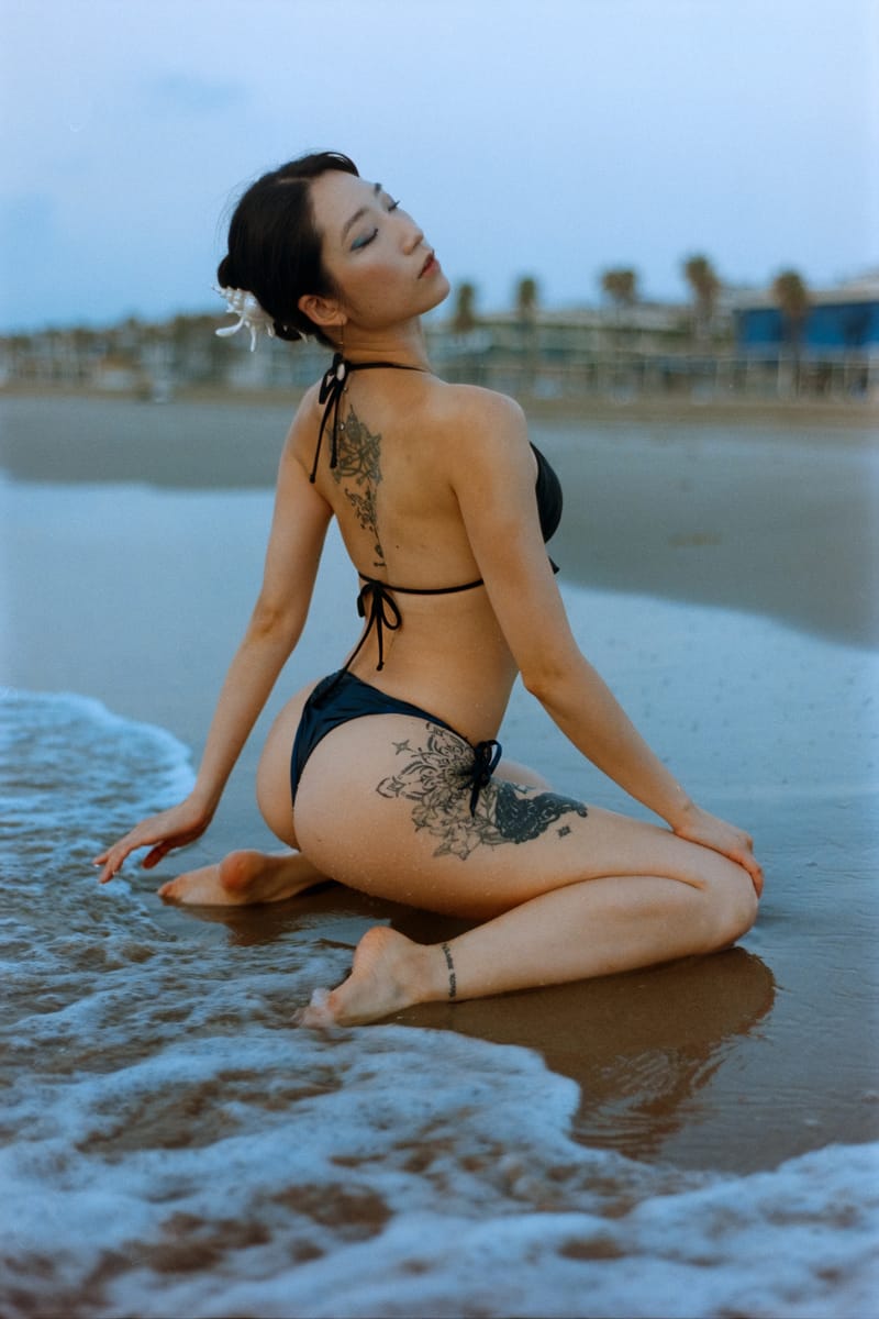 A woman in a black bikini sits on wet sand at the shoreline, back arched toward the camera, floral tattoos across her spine and thigh, with a blurred palm-lined boardwalk and soft dusk light behind her.